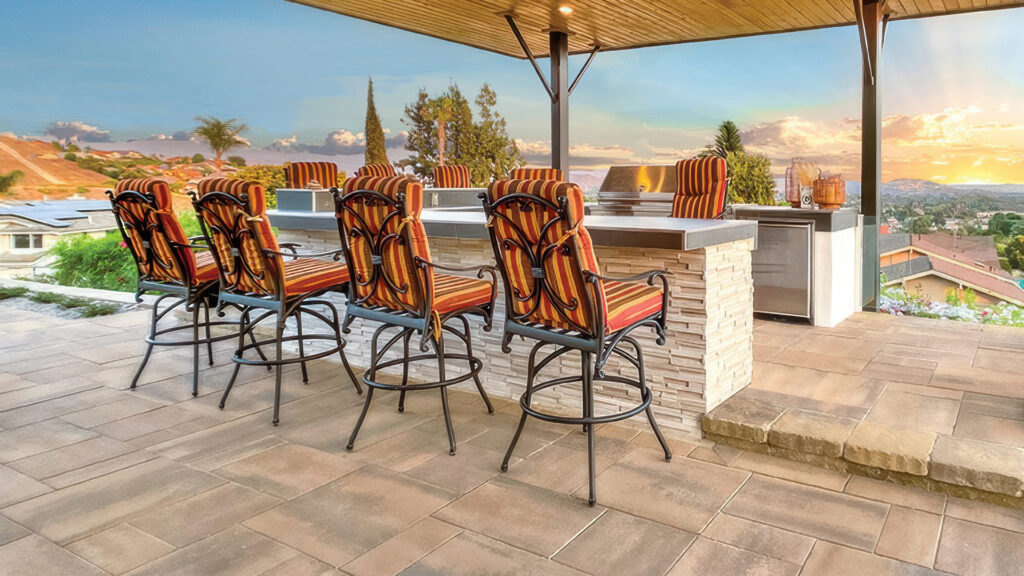 Covered outdoor kitchen and dining area featuring Paseo I & II pavers in Adobe-Copper-Mocha installed in an ashlar pattern. A step down to a lower patio is finished with tumbled Sand-Stone-Mocha Estate Cobble II. Bar seating includes striped cushioned chairs at a built-in BBQ island with stacked stone veneer and stainless steel appliances.