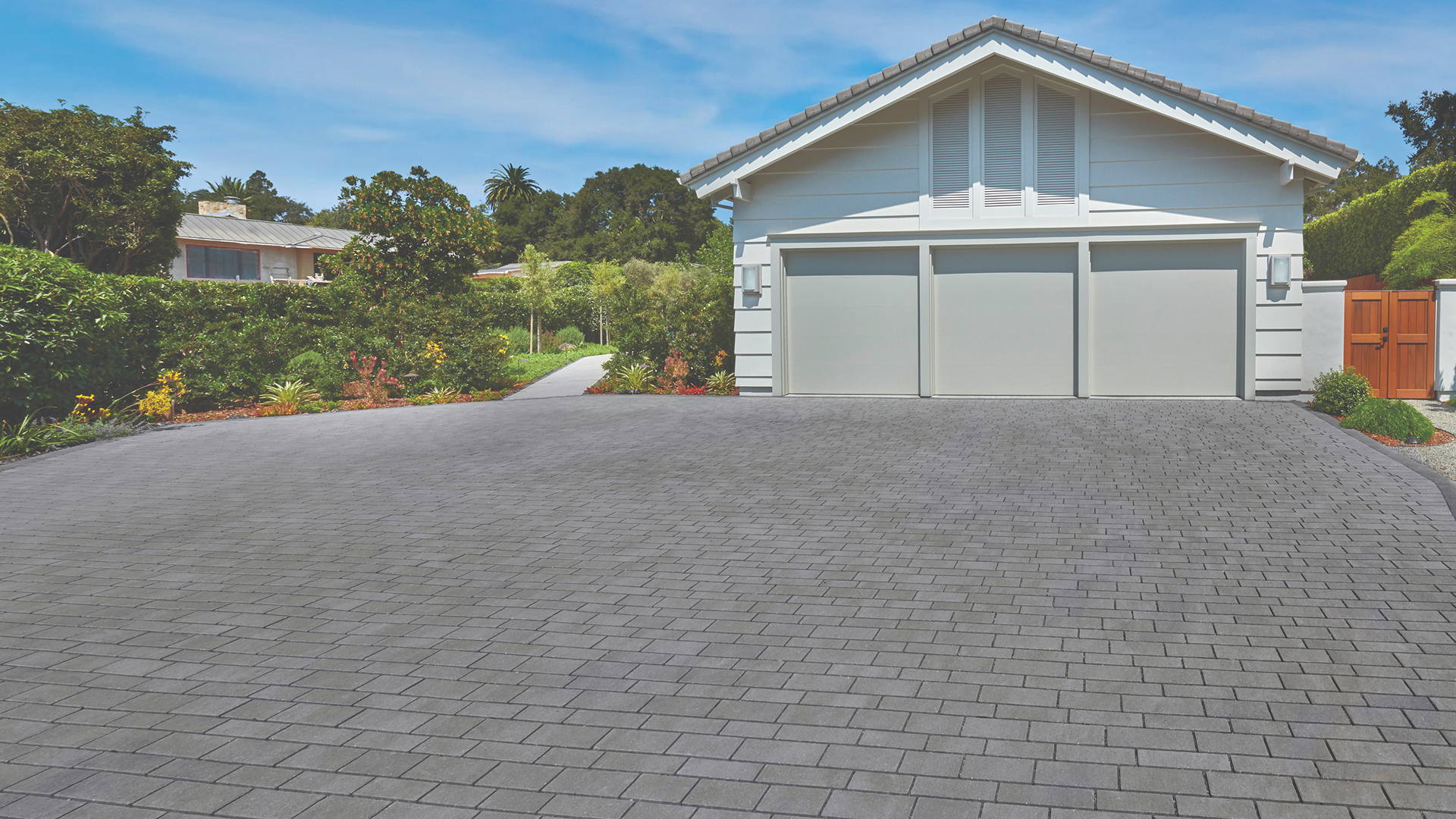 Driveway paved with Dark Gray-Pewter-Charcoal Permeable Aqualina pavers in a random runner pattern, leading up to a modern white three-car garage surrounded by lush landscaping.