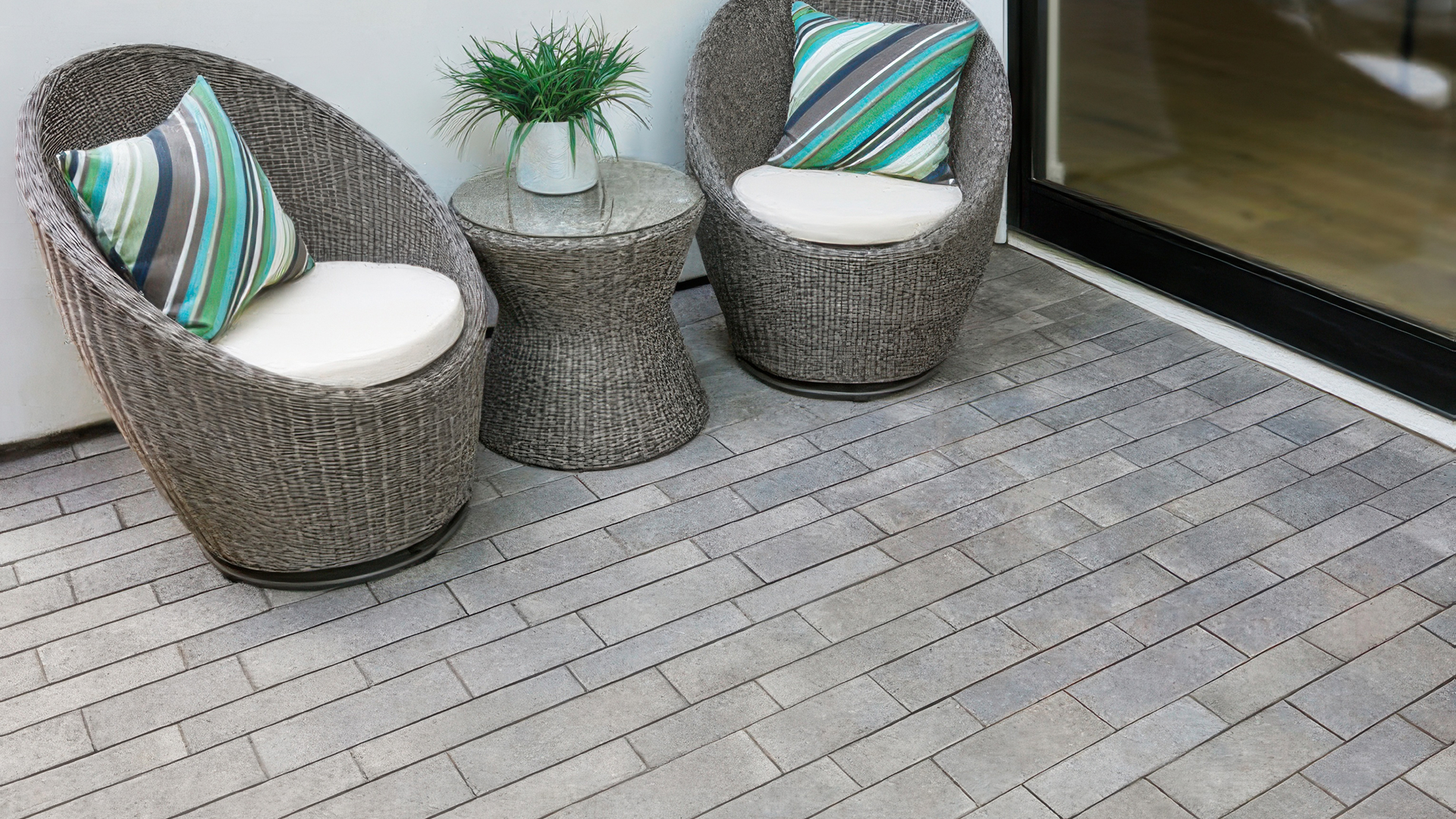 Cozy outdoor patio area featuring Dark Gray-Pewter-Charcoal Aqualina pavers installed in a random runner pattern. The space includes two round wicker chairs with striped accent pillows and a matching side table with a small plant, set against a modern white wall and glass door.