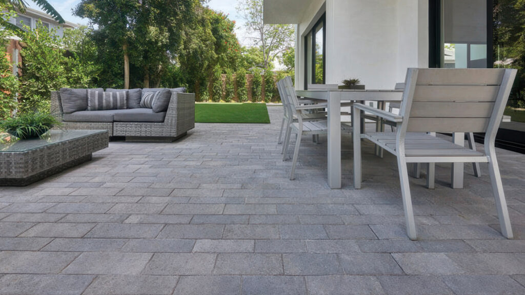 Modern outdoor patio featuring Dark Gray-Pewter-Charcoal Aqualina pavers installed in a random runner pattern. The clean-lined pavers create a sleek, contemporary surface beneath a white dining set and gray outdoor sofa, surrounded by lush greenery.