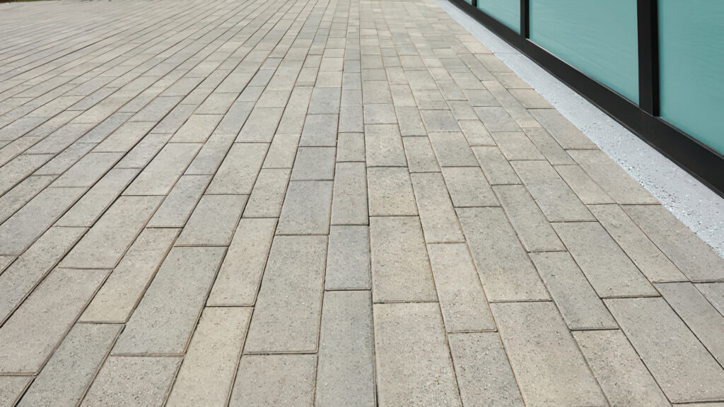 Close-up of Aqualina paving stones in Dark Gray–Pewter–Charcoal blend, installed in a random runner pattern. The modern, linear layout is placed alongside a contemporary building with frosted glass panels and a concrete edge.