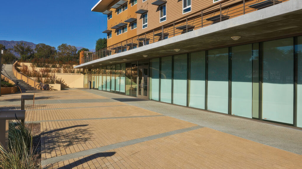 Modern university or office building with a wide pedestrian plaza paved in Sand Permeable Holland pavers, installed in a running bond pattern. The walkway features subtle linear accents in a contrasting color and is bordered by glass windows and landscaped stairways. The setting is framed by distant mountains under a clear blue sky.