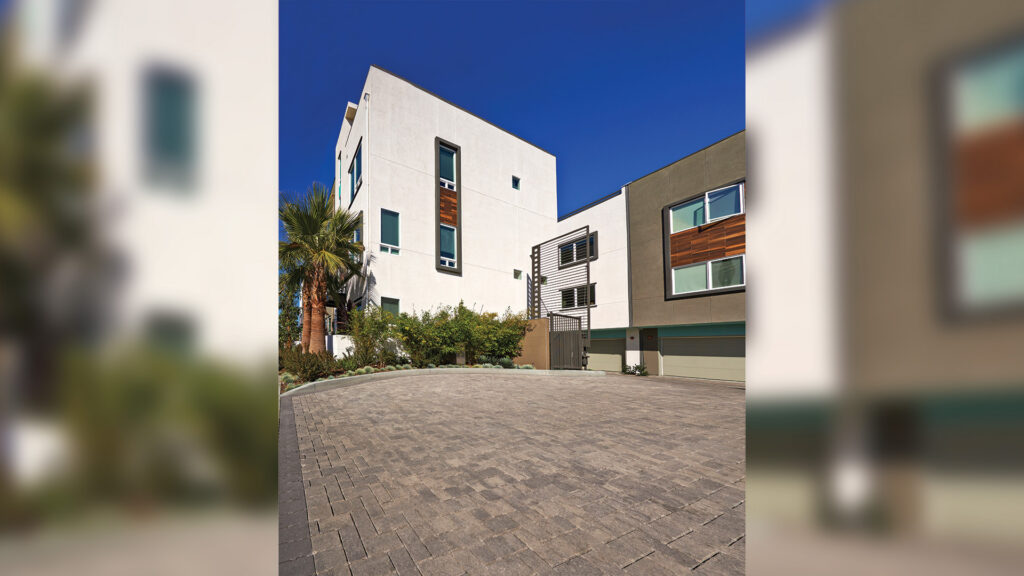 Driveway of a contemporary townhouse complex paved with 80mm Permeable Holland pavers in a Gray-Charcoal blend, arranged in a herringbone pattern. The driveway is bordered with solid Charcoal Holland pavers in a soldier course. Modern architecture with white and earth-toned facades, wood panel accents, and a gated entrance enhance the clean, structured look under a deep blue sky.