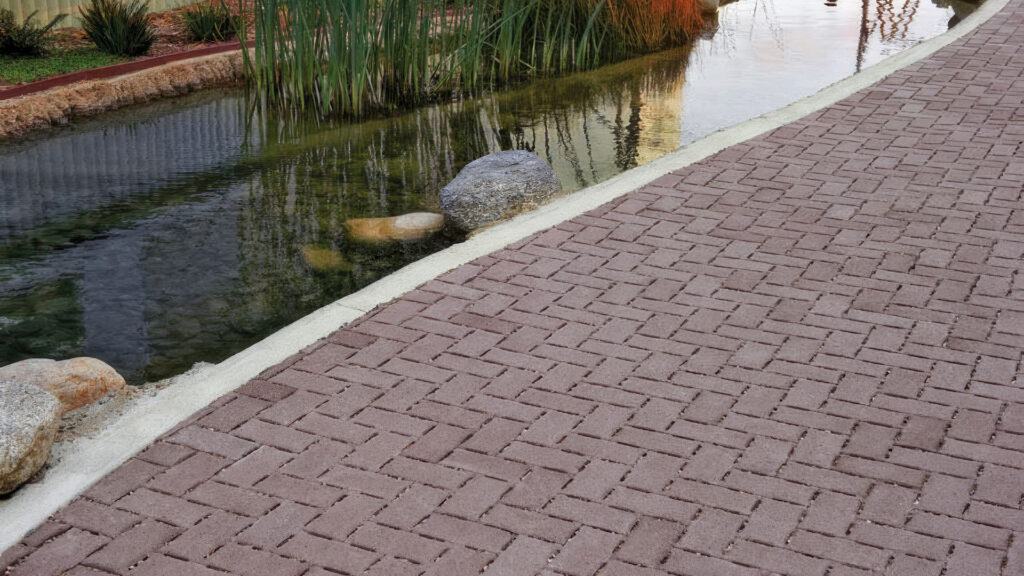 Curved pathway paved with Permeable Holland pavers in a special order Brown color, arranged in a herringbone pattern. The path runs alongside a narrow stream with natural rocks, aquatic plants, and gentle ripples reflecting nearby landscaping and architecture. The scene blends functional paving with serene, nature-inspired design.