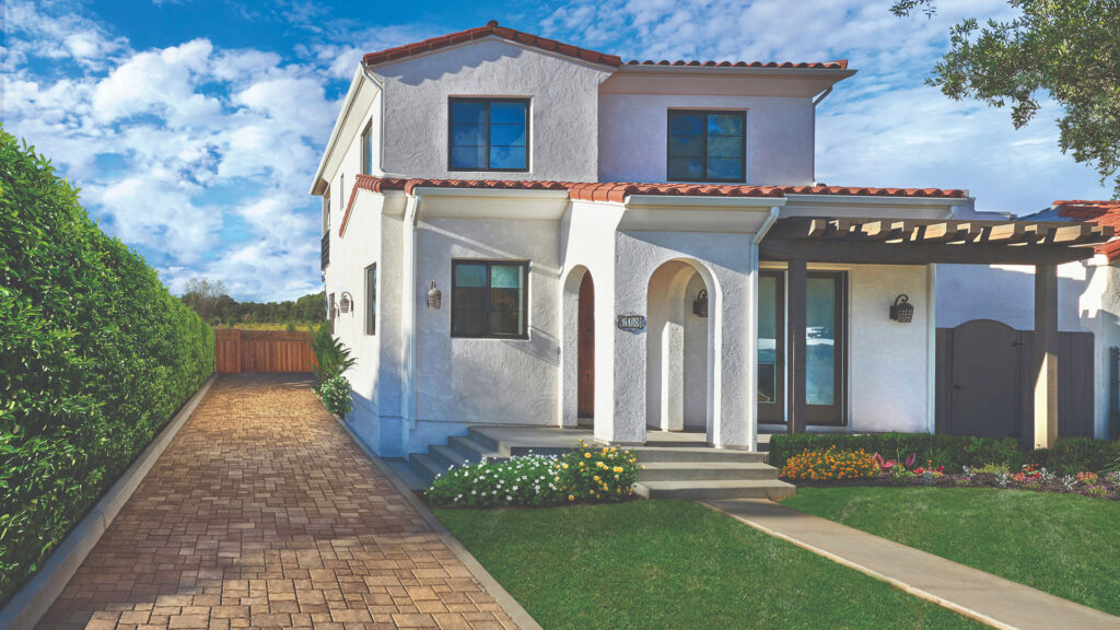 Side driveway paved with Sand-Stone-Mocha Permeable Slate in a random ashlar pattern, leading alongside a white stucco home with terracotta roof tiles, surrounded by lush greenery and vibrant flower beds.