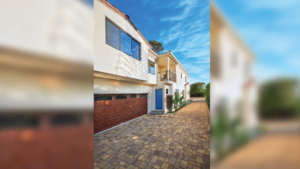 Driveway paved with Sand-Stone-Mocha Permeable Slate in a random ashlar pattern, leading to a two-story stucco home with a blue front door and wooden garage doors under a bright blue sky.