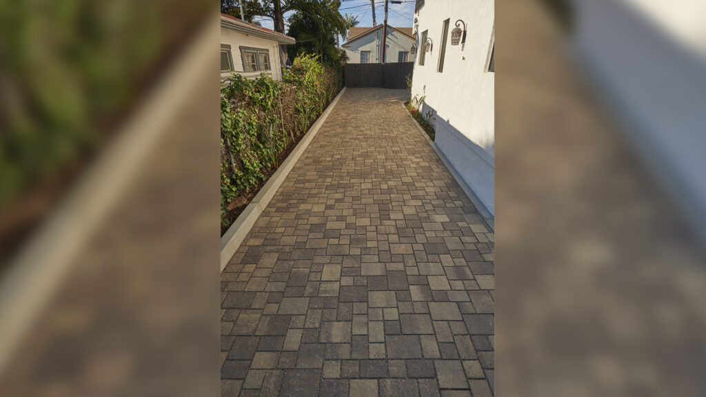 Side driveway paved with Sand-Stone-Mocha Permeable Slate in a random ashlar pattern, bordered by concrete curbs and adjacent to a white stucco house and garden hedge.