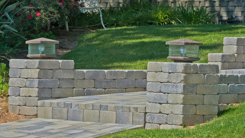 Garden pathway featuring columns built from Gray-Moss-Charcoal Rustic Wall blocks, accented with decorative lantern lights. The pathway and steps are paved with Sand-Stone-Mocha Courtyard Stone in a neat, modular layout, complementing the lush green lawn and vibrant flower beds in the background.
