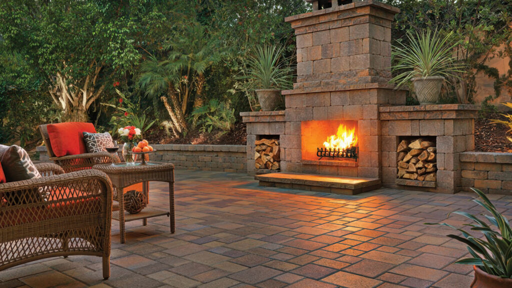 Outdoor patio scene featuring a Tuscan Rustic Wall fireplace and seating wall. The fireplace includes wood storage and is constructed with field-split Courtyard Stone pavers and Rustic Wall elements. The patio surface is paved in a 50/50 blend of Sand-Stone-Mocha Courtyard Stone pavers arranged in an ashlar pattern. Wicker lounge furniture with red cushions and a side table add a cozy touch, surrounded by lush greenery and soft lighting.