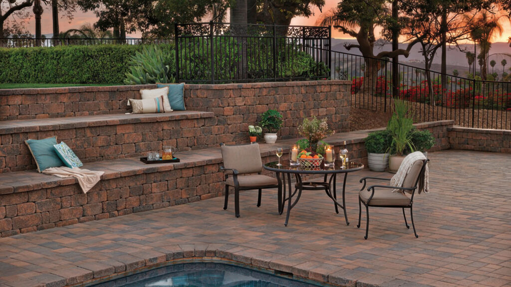 Elegant poolside patio at sunset featuring Stonewall Block and Cap in Cream-Brown-Charcoal used for the terraced seating walls and pool coping. The paving is done with Courtyard Combo Stone in Cream-Brown-Charcoal, laid in a random ashlar pattern. Cushions, potted plants, and a round dining table set with candles and drinks create a warm, inviting outdoor living space with a scenic view.