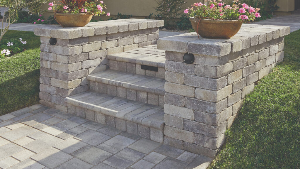 Outdoor entry steps flanked by Gray-Moss-Charcoal Rustic Wall Stone used to build low walls with decorative lighting fixtures. The steps are surfaced with Gray-Charcoal Bullnose for a smooth, rounded edge, while the surrounding patio is paved with Gray-Charcoal Courtyard Combo stone laid in an ashlar pattern. Two large planters with pink flowers sit atop the wall columns, adding a vibrant touch to the soft-toned stonework.