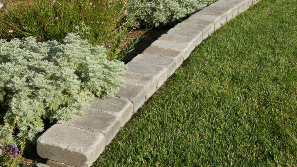 Raised curb made from Rustic Wall in Gray-Moss-Charcoal color, forming a clean edge between a green lawn and a lush, flowering plant bed.