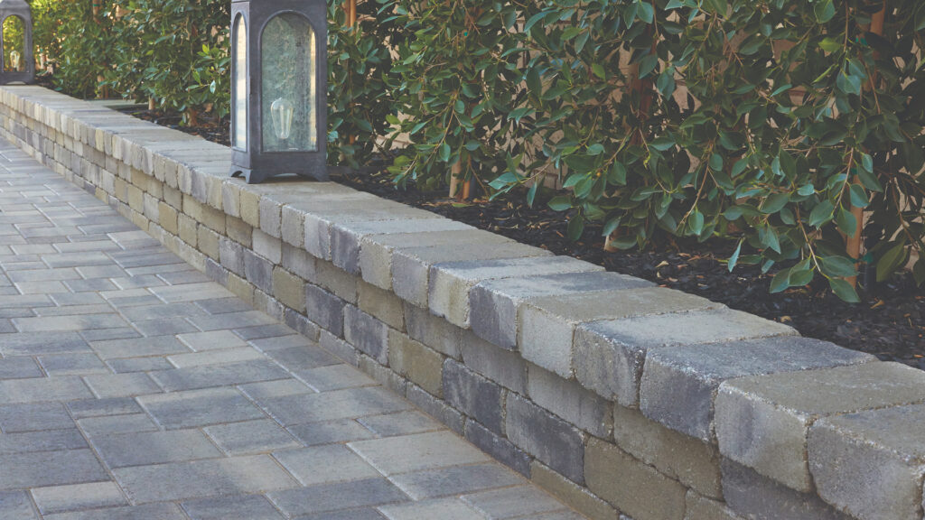 Gray-Moss-Charcoal Rustic Wall planter wall borders a walkway lined with lush greenery. The wall is topped with smooth stone caps and decorative lanterns. The paving surface features Gray-Charcoal Courtyard Combo pavers installed in a random ashlar pattern, creating a refined and structured look.