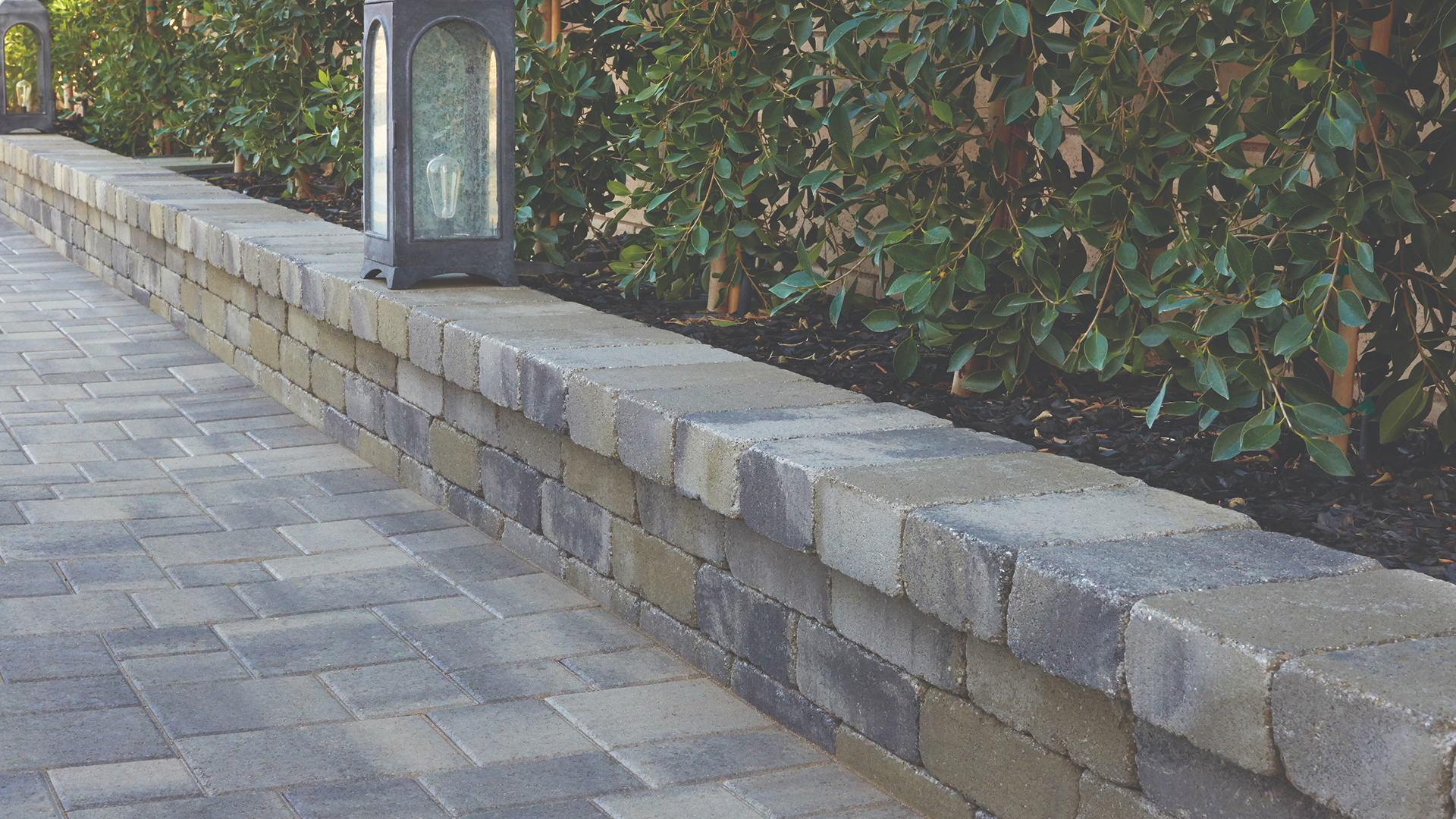Gray-Moss-Charcoal Rustic Wall planter wall borders a walkway lined with lush greenery. The wall is topped with smooth stone caps and decorative lanterns. The paving surface features Gray-Charcoal Courtyard Combo pavers installed in a random ashlar pattern, creating a refined and structured look.