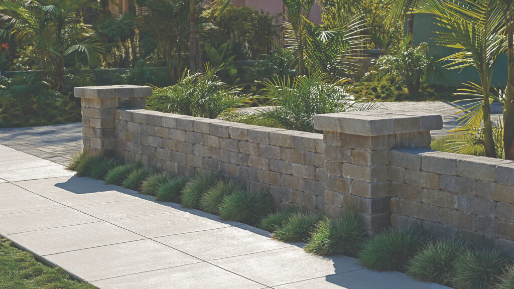Gray-Moss-Charcoal Rustic Wall Stone used to build a low retaining wall with decorative columns, bordering a landscaped yard with ornamental grasses and palm plants alongside a clean, modern sidewalk.