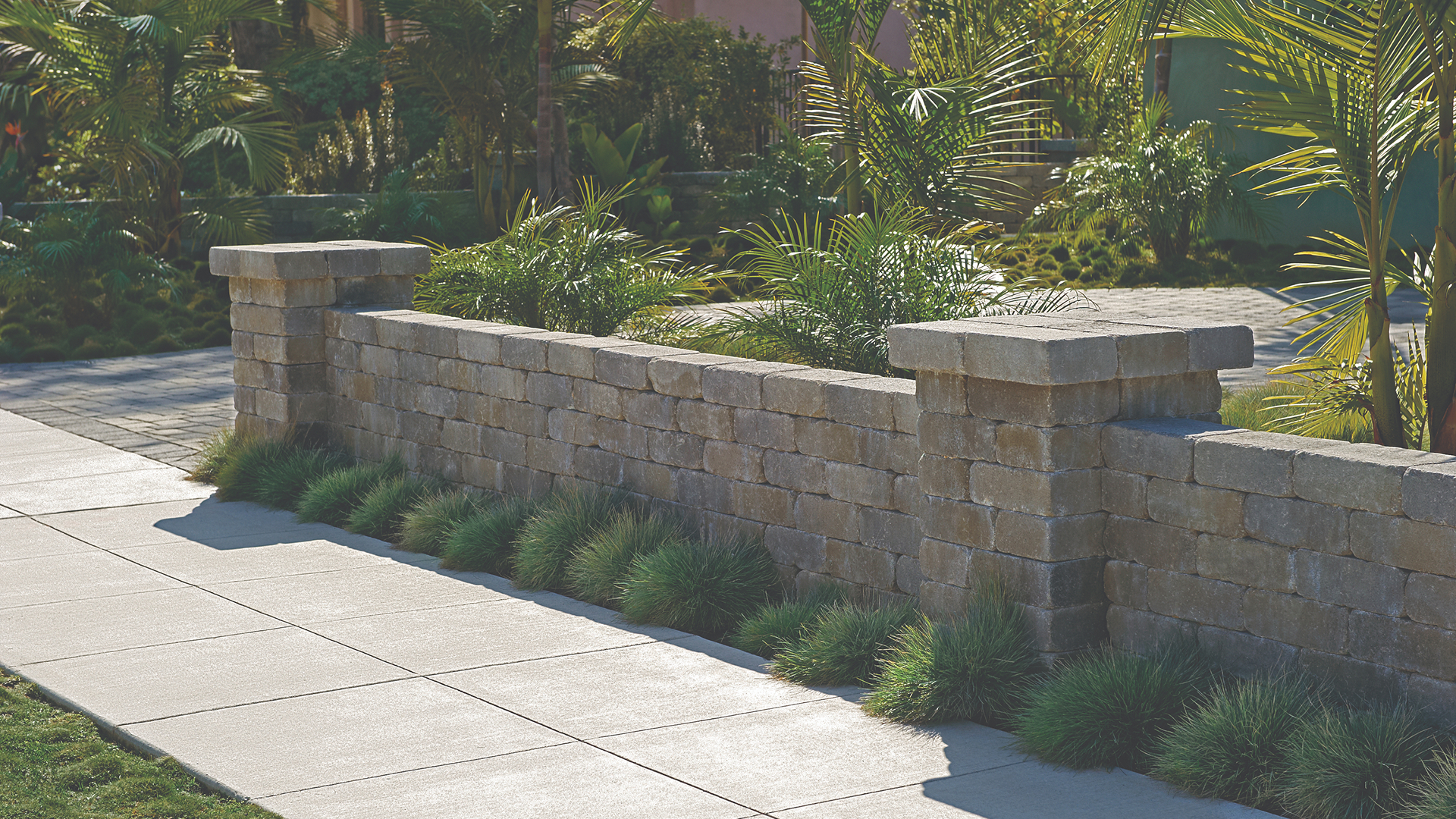 Gray-Moss-Charcoal Rustic Wall Stone used to build a low retaining wall with decorative columns, bordering a landscaped yard with ornamental grasses and palm plants alongside a clean, modern sidewalk.