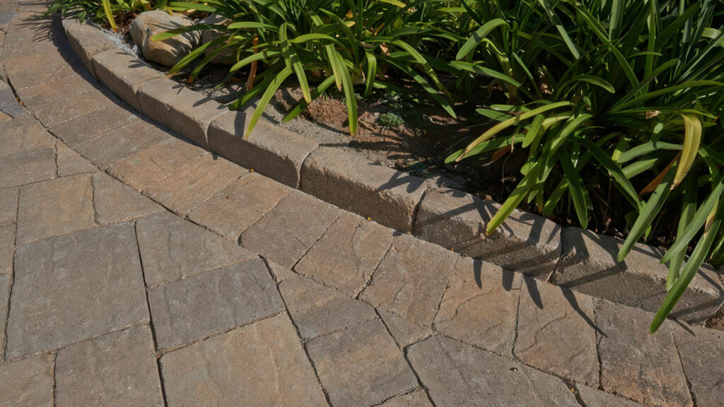 Detail view of a raised curb constructed with Rustic Wall units in Sand-Stone-Mocha. The paving surface features a 50/50 blend of Cream-Brown-Charcoal and Sand-Stone-Mocha Slate Stone Combo (distressed) in an ashlar pattern. Border edging is Slate Stone 6x9 (distressed) in Cream-Brown-Charcoal.