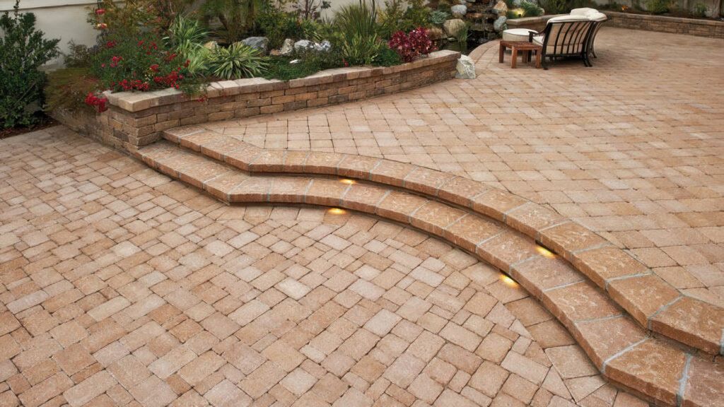 Outdoor patio featuring Sand-Stone Castle Cobble I & II pavers with a tumbled finish, installed in a combination of random ashlar and stack bond patterns. The space includes curved steps with in-step lighting and a low garden wall, creating a warm, inviting landscape design.