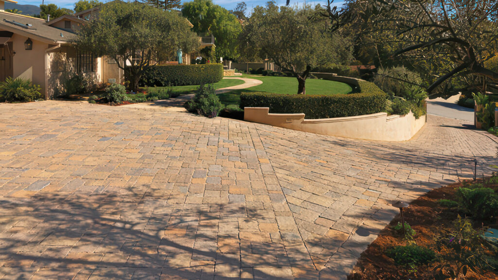 Residential driveway paved with a 50/50 blend of Sand-Stone-Mocha and Tuscan Estate Cobble I & II, installed in an ashlar pattern. The driveway slopes gently and is surrounded by manicured hedges, grass, and mature trees in a lush landscape.