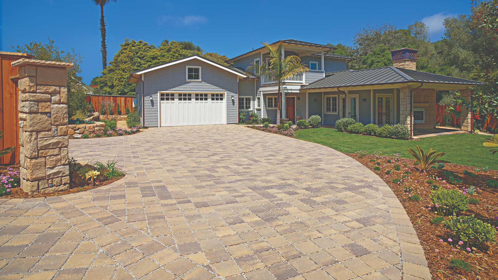 Driveway paved with SF Rima pavers in Sand–Stone–Mocha blend, installed in a stack bond pattern. The setting features a coastal-style home with white garage doors, light gray siding, and vibrant landscaping with flowering plants and neatly trimmed grass.