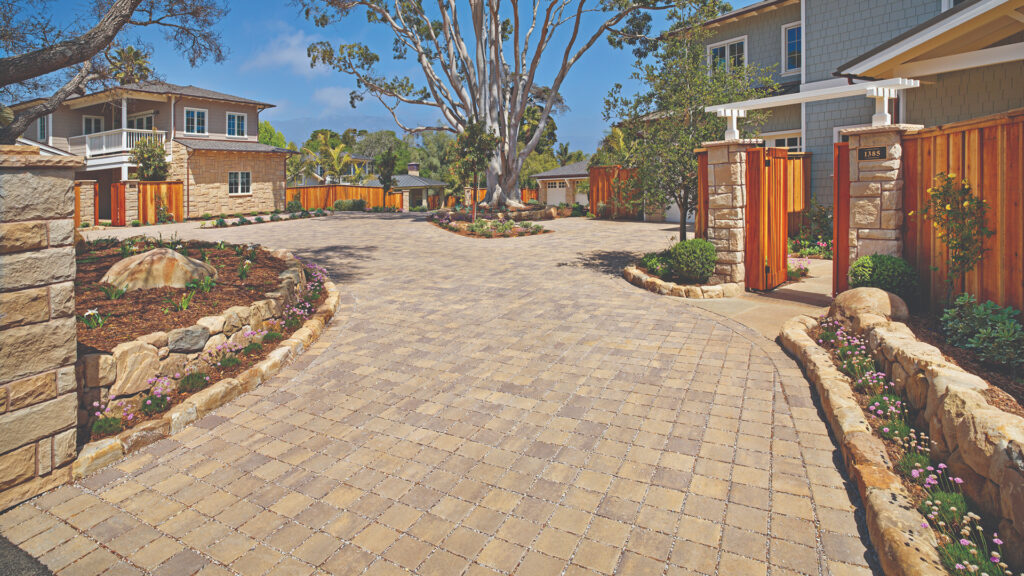 Shared residential driveway paved with SF Rima pavers in Sand–Stone–Mocha blend, installed in a stack bond pattern. The setting features multiple Craftsman-style homes, lush landscaping, natural stone borders, and a large central tree as a focal point.
