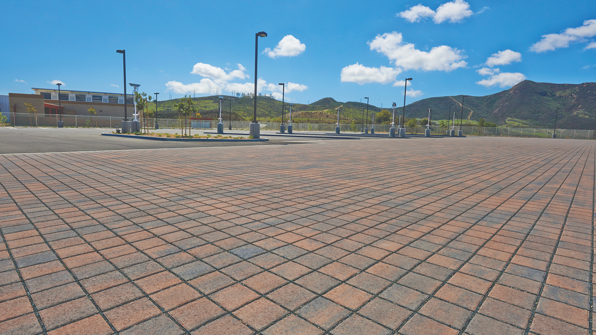 Expansive parking area paved with Cream-Charcoal SF Rima pavers in a stack bond pattern. The permeable pavers create a uniform grid appearance, framed by light poles and a nearby school building. Rolling green hills and a bright blue sky with scattered clouds form the scenic backdrop.
