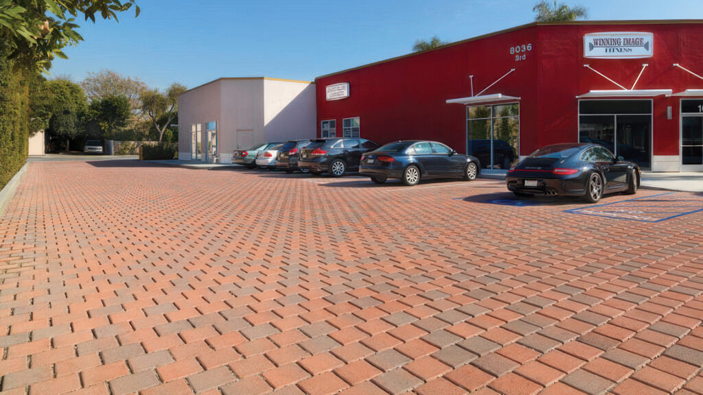 Commercial parking lot paved with Red-Brown SF Rima (special order color) in a running bond pattern. Multiple cars are parked in front of a bright red building with white signage and glass storefronts. The open lot is bordered by hedges and trees under a clear blue sky.
