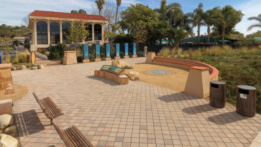Public park plaza paved with Cream-Charcoal SF Rima (special order color) pavers in a 45-degree angled stack bond pattern. A decorative circular tile mosaic is featured in a sandy section surrounded by curved seating. The space includes interpretive signage, benches, and natural landscaping with native grasses and trees. A Spanish-style building with red tile roofing is visible in the background.