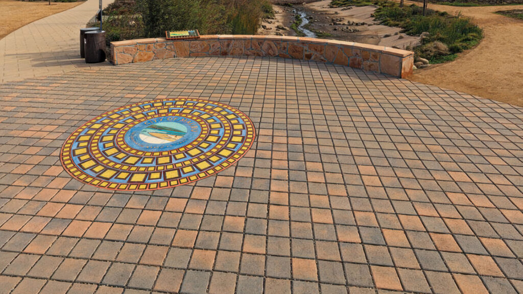 Public plaza featuring Cream-Charcoal SF Rima pavers (special order color) installed in a 45-degree angled stack bond pattern. A vibrant circular tile mosaic inset in the center displays the seal of the City of Aliso Viejo with blue, yellow, and red geometric rings. The space is framed by a low stone wall and overlooks a landscaped creek area with walking paths.