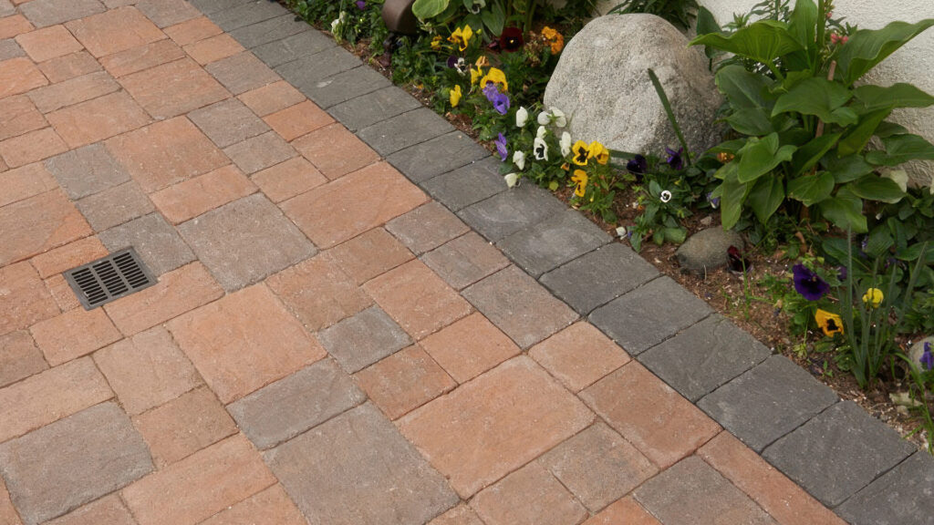Drains & Caps: 50/50 blend of Cream-Terracotta-Brown and Cream-Brown-Charcoal distressed Slate Stone Combo installed in an ashlar pattern. A solid Charcoal 6x9 distressed Slate Stone border defines the edge next to a planted garden bed, with a square metal drain integrated into the paving.