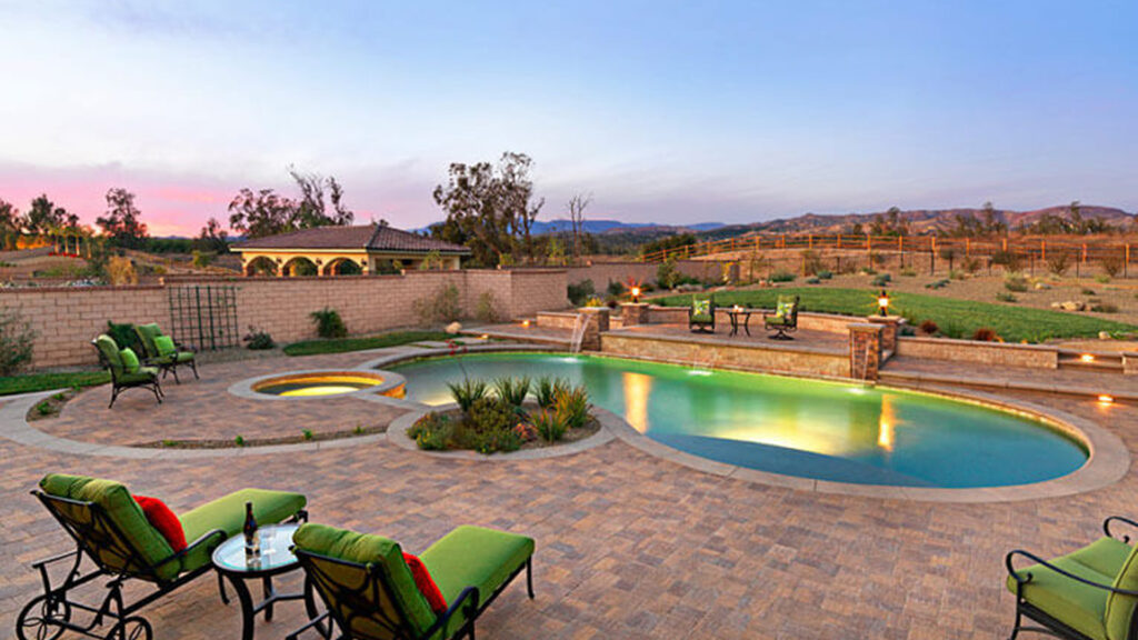 Backyard oasis featuring Slate Stone pavers installed in an ashlar pattern surrounding a freeform pool and spa. The space includes cushioned lounge chairs, a raised water feature with cascading spouts, and soft lighting throughout. Desert landscaping and distant hills complete the serene sunset setting.