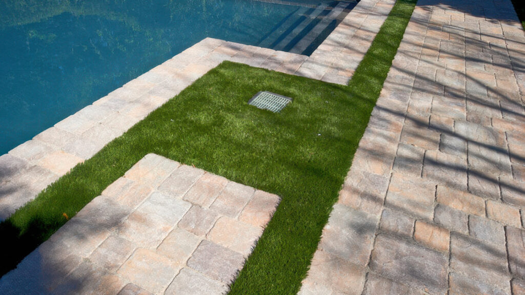 Poolside area featuring Tuscan tumbled Slate Stone Combo pavers installed in an ashlar pattern with grass inlays and a centered drain. Pool coping is completed with Tuscan Slate Stone 6x9 pavers in a tumbled finish for a cohesive, natural look.