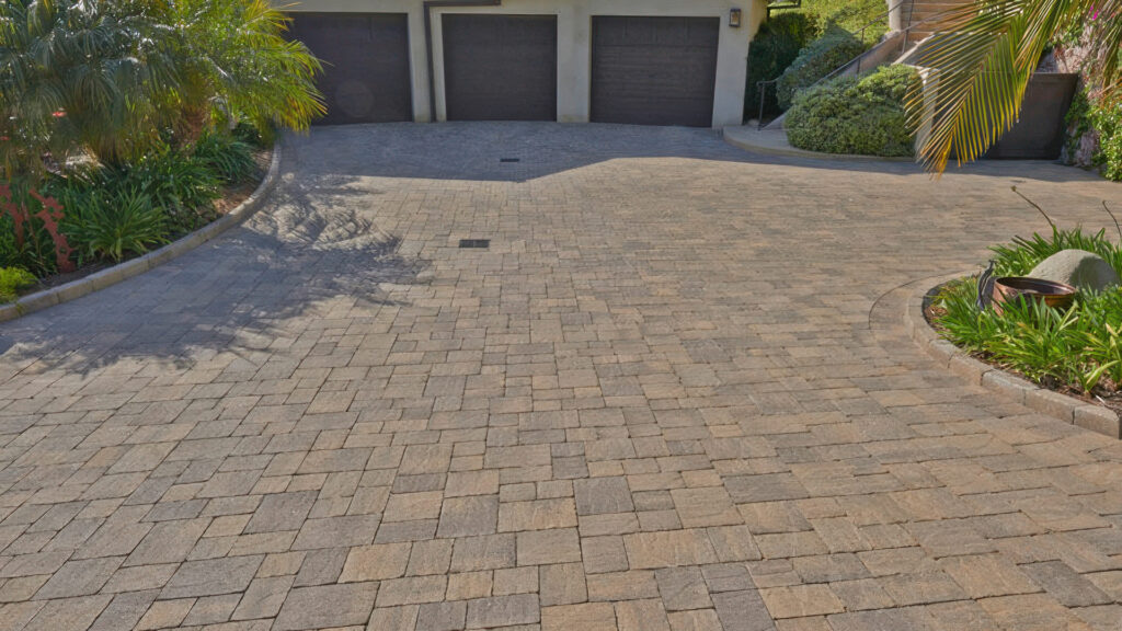 Driveway installation features a 50/50 blend of Sand-Stone-Mocha and Cream-Brown-Charcoal Slate Stone Combo (distressed), arranged in an ashlar pattern for a natural, textured look. The surface is bordered by soft landscaping and leads to a garage.