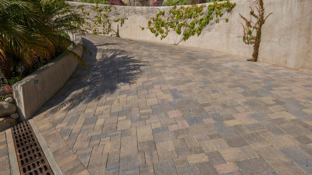 Driveway paved with a 50/50 blend of Sand-Stone-Mocha and Cream-Brown-Charcoal distressed Slate Stone Combo in an ashlar pattern. The surface features a subtle mix of earth tones with a linear drain integrated at the base of the slope for water management.