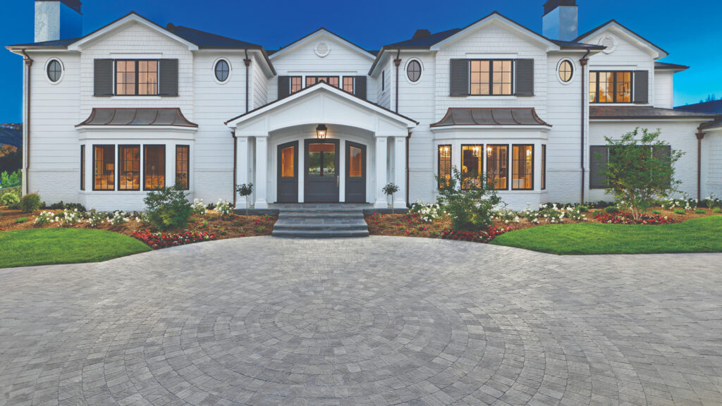 Grand entrance with Slate Stone Circle package in Gray–Charcoal blend centered in the driveway, surrounded by Slate Stone Combo pavers in Gray–Charcoal installed in a random ashlar pattern. The setting features a white colonial-style home with manicured lawns and colorful flower beds.