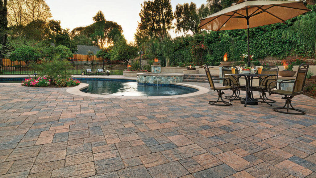 Backyard patio surrounding a curved-edge pool, featuring Cream-Brown-Charcoal Slate Stone pavers installed in an ashlar pattern. The area includes a round dining table with chairs under a large umbrella, lush landscaping with pink flowers, and multiple fire features in the background, creating a warm and inviting atmosphere at sunset.
