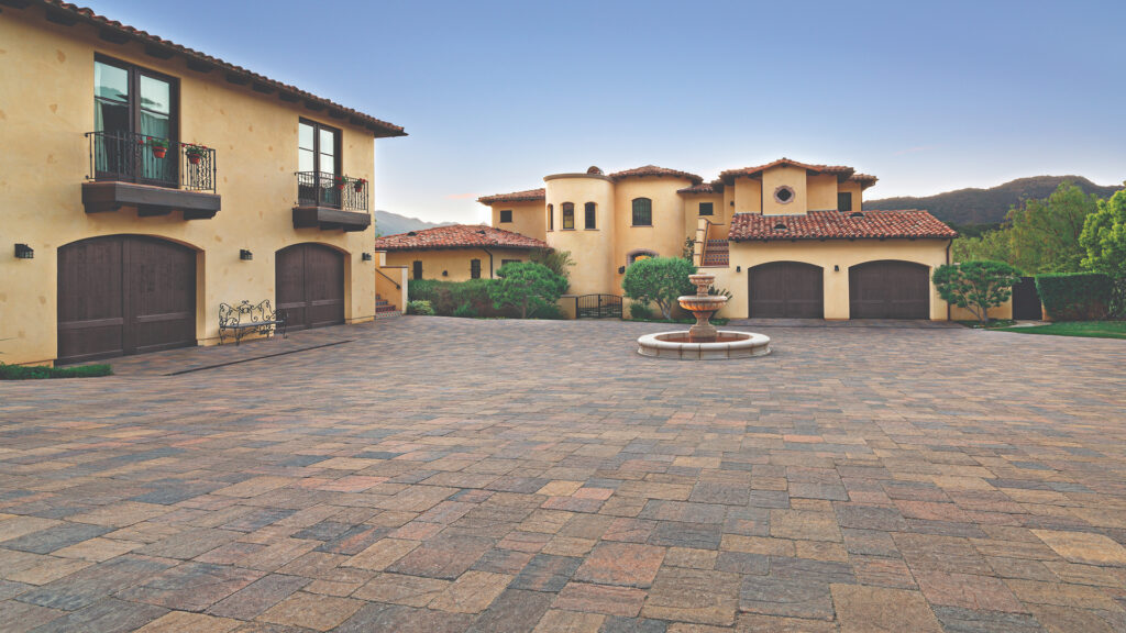Spacious courtyard paved with a 50/50 field mix of Slate Stone in Sand–Stone–Mocha and Cream–Brown–Charcoal blends, installed in a random ashlar pattern. The setting features a Mediterranean-style estate with warm stucco walls, terracotta roof tiles, and a central water fountain.