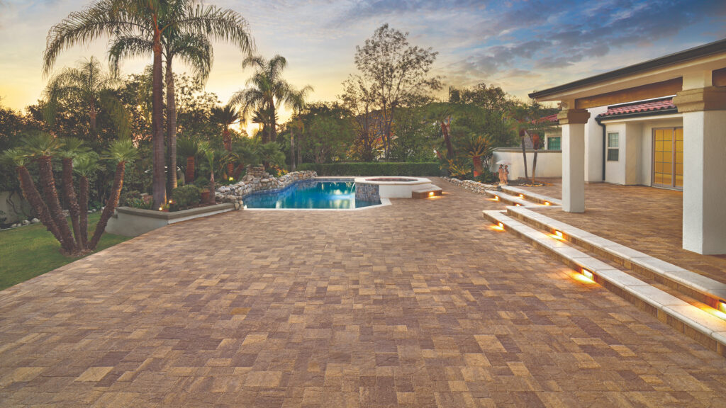 Expansive tropical-style backyard featuring Sand-Stone-Mocha Slate Stone pavers installed in a random ashlar pattern. The area includes a freeform pool with built-in spa, tiered steps with integrated lighting, and lush landscaping with palm trees and rock features, all set against a dramatic sunset sky.