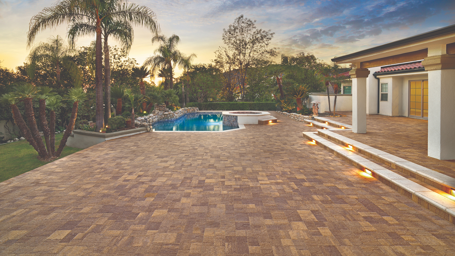 Expansive tropical-style backyard featuring Sand-Stone-Mocha Slate Stone pavers installed in a random ashlar pattern. The area includes a freeform pool with built-in spa, tiered steps with integrated lighting, and lush landscaping with palm trees and rock features, all set against a dramatic sunset sky.