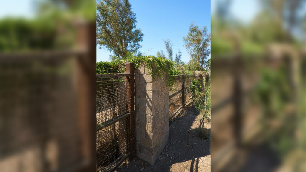 Outdoor fencing with integrated column constructed using Sand-Stone-Mocha Stonewall II block and cap. The sturdy column supports a metal mesh gate and is topped with greenery cascading over the edge, blending function with natural aesthetics in a rural or garden setting.
