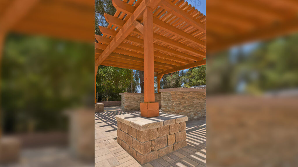 Outdoor patio area featuring a wooden patio cover supported by a base column made from Sand-Stone-Mocha Stonewall II block and cap. The ground is paved with Sand-Stone-Mocha Courtyard Stone pavers in a clean, structured layout that complements the warm tones of the wooden structure and hardscape.