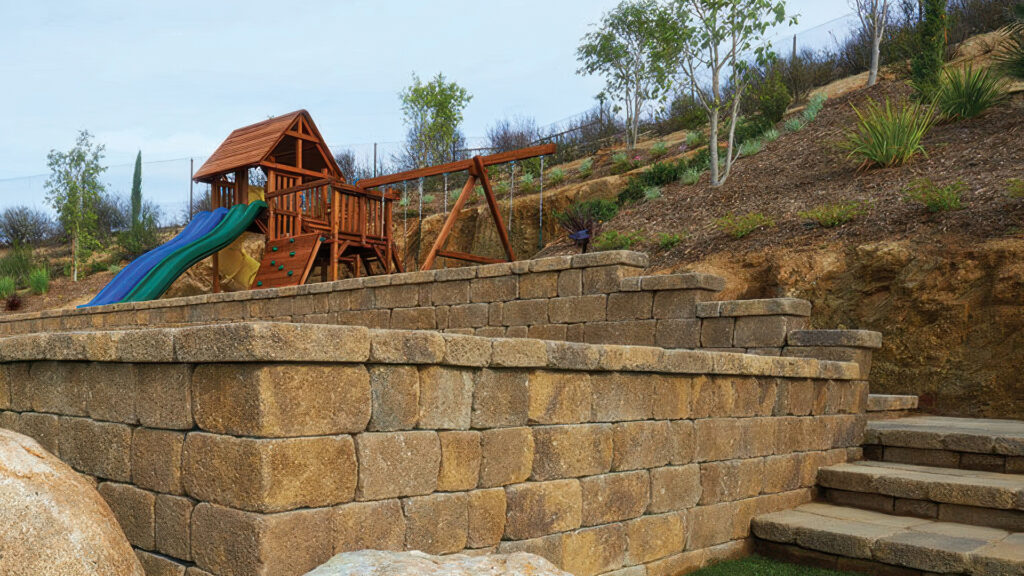 Multi-level retaining wall built with Sand-Stone-Mocha Stonewall® II block and cap, supporting a terraced hillside landscape. A wooden playground structure with swings and slides is placed on the top level, surrounded by young trees and hillside plantings. Stone steps lead up alongside the wall.