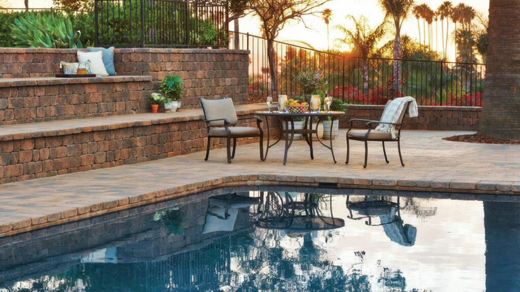 Elegant poolside patio at sunset featuring Cream-Brown-Charcoal Stonewall® II used for multi-level seating walls and pool coping. The patio surface is paved with Cream-Brown-Charcoal Courtyard Stone laid in an ashlar pattern. A round dining table with drinks and fruit, cushioned chairs, and potted plants create a warm and inviting outdoor setting surrounded by lush greenery and a backdrop of palm trees.