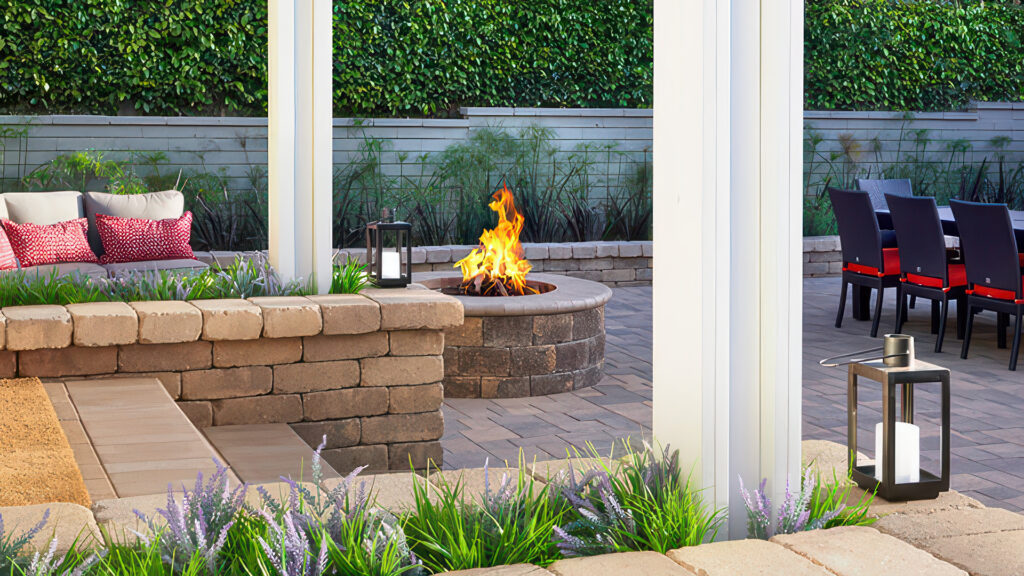 Outdoor patio featuring a Sand-Stone-Mocha Stonewall® Fire Pit kit capped with Bullnose in solid Stone color, surrounded by Courtyard Combo pavers in Sand-Stone-Mocha laid in a random ashlar pattern. A planter wall in the background is built with Sand-Stone-Mocha Rustic wall blocks, creating a cozy seating and dining area with modern furnishings and lush landscaping.