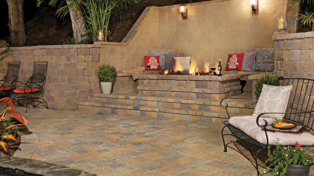 Cozy nighttime patio scene featuring a built-in fire pit and seating area made with Tuscan Stonewall® II blocks. The surrounding walls and steps are constructed from the same material. The patio surface and steps are paved with a 50/50 blend of Sand-Stone-Mocha and Gray-Moss-Charcoal Slate Stone in a random ashlar pattern. Cushioned seating, decorative pillows, and ambient lighting enhance the warm, inviting atmosphere.