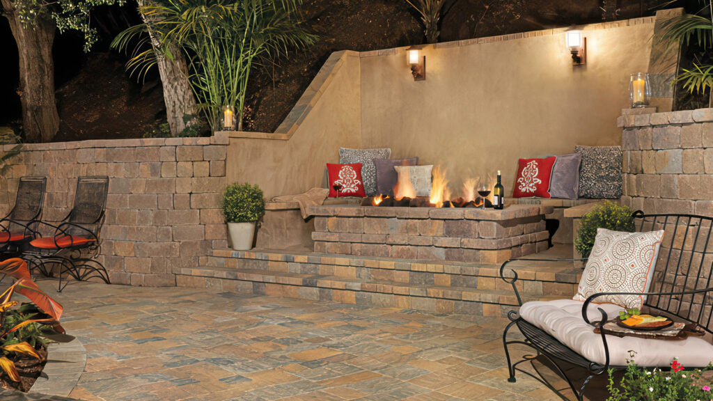 Cozy outdoor seating area with a built-in firepit and bench-style seat made from Stonewall II block and cap in Tuscan color. The retaining wall surrounding the space is also constructed from matching Stonewall II material. The patio flooring features Slate Stone pavers (distressed) in an ashlar pattern, with a 50/50 blend of Sand-Stone-Mocha and Gray-Moss-Charcoal colors, creating a rich and textured surface.
