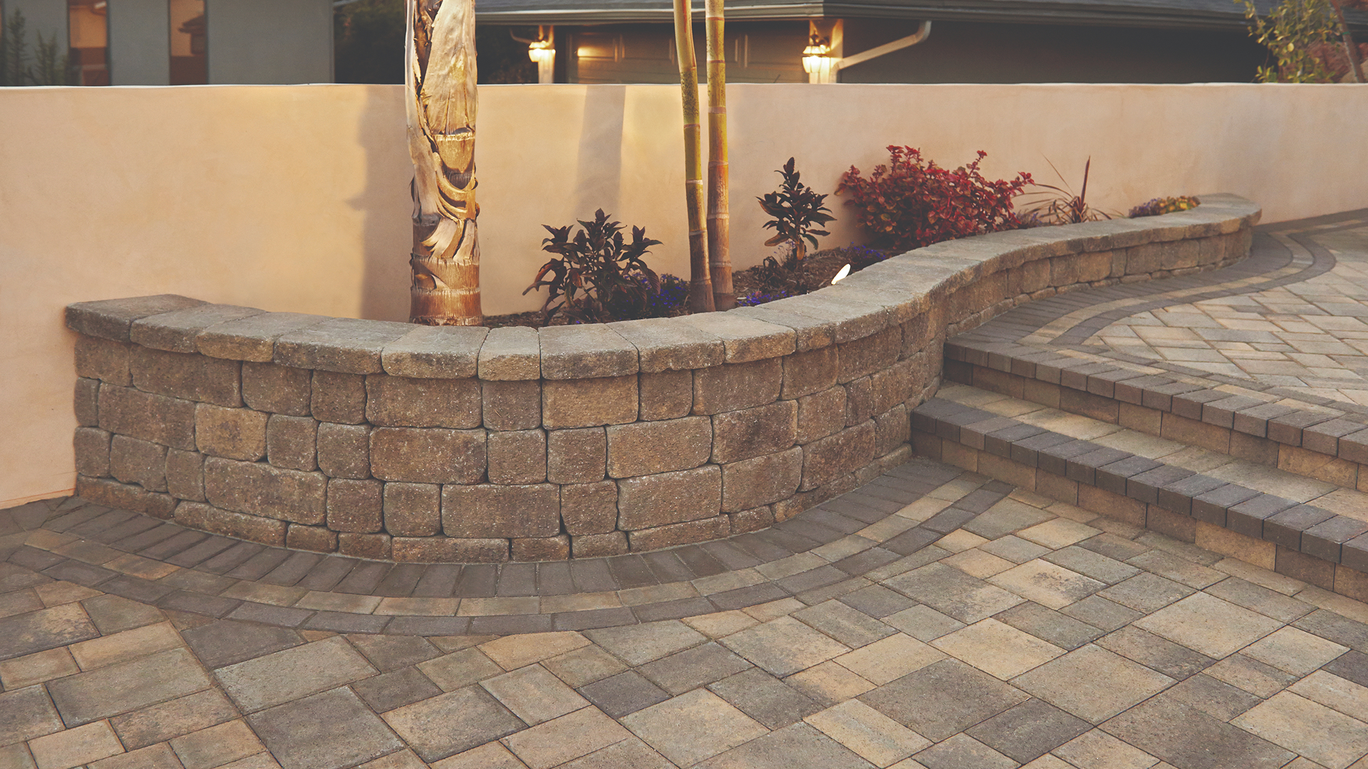 Curved planter wall constructed with Sand-Stone-Mocha Stonewall® II block and cap. The patio surface is paved with Courtyard Combo pavers in Sand-Stone-Mocha laid in a random ashlar pattern. Decorative border detail includes a soldier course of Holland Mocha, followed by Holland Sand-Stone-Mocha and Holland Mocha in running bond. Steps lead to a raised level with additional planting, framed by soft lighting and tropical foliage.