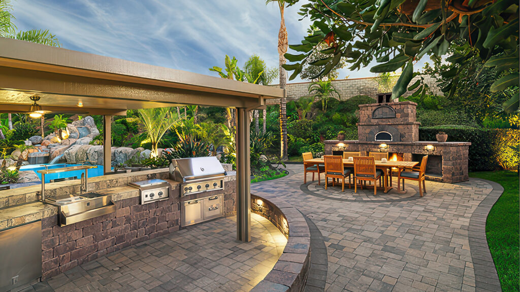 Luxurious backyard kitchen and dining area featuring a custom-built Stonewall® II BBQ island and fireplace in Tuscan. The patio is paved with Slate Stone in Dark Gray-Copper-Charcoal (special order) in a random ashlar pattern. Accenting the space are border and banding details in solid Charcoal Slate 6x9 pavers, with a decorative circular inlay in Slate Circle Gray-Charcoal. Surrounded by lush tropical landscaping and ambient lighting.