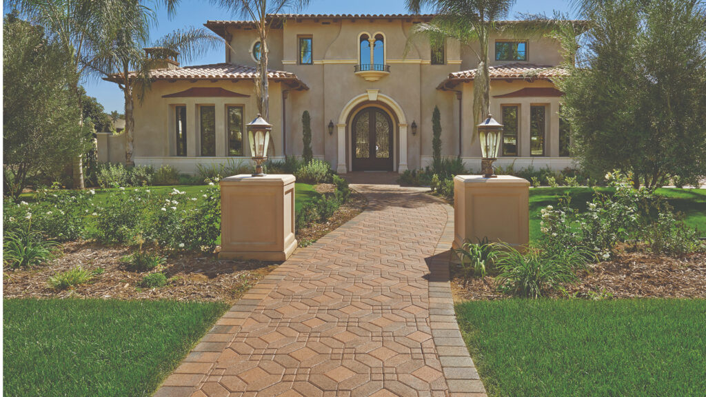 Cream-Brown Symetry® paving stones (special order color) combined with solid Cream 6x6 squares arranged in a Venetian parquet pattern. The elegant curved walkway is bordered with Tuscan Antique Cobble I and leads to a grand front entrance of a Mediterranean-style home framed by manicured landscaping and decorative lamp posts.