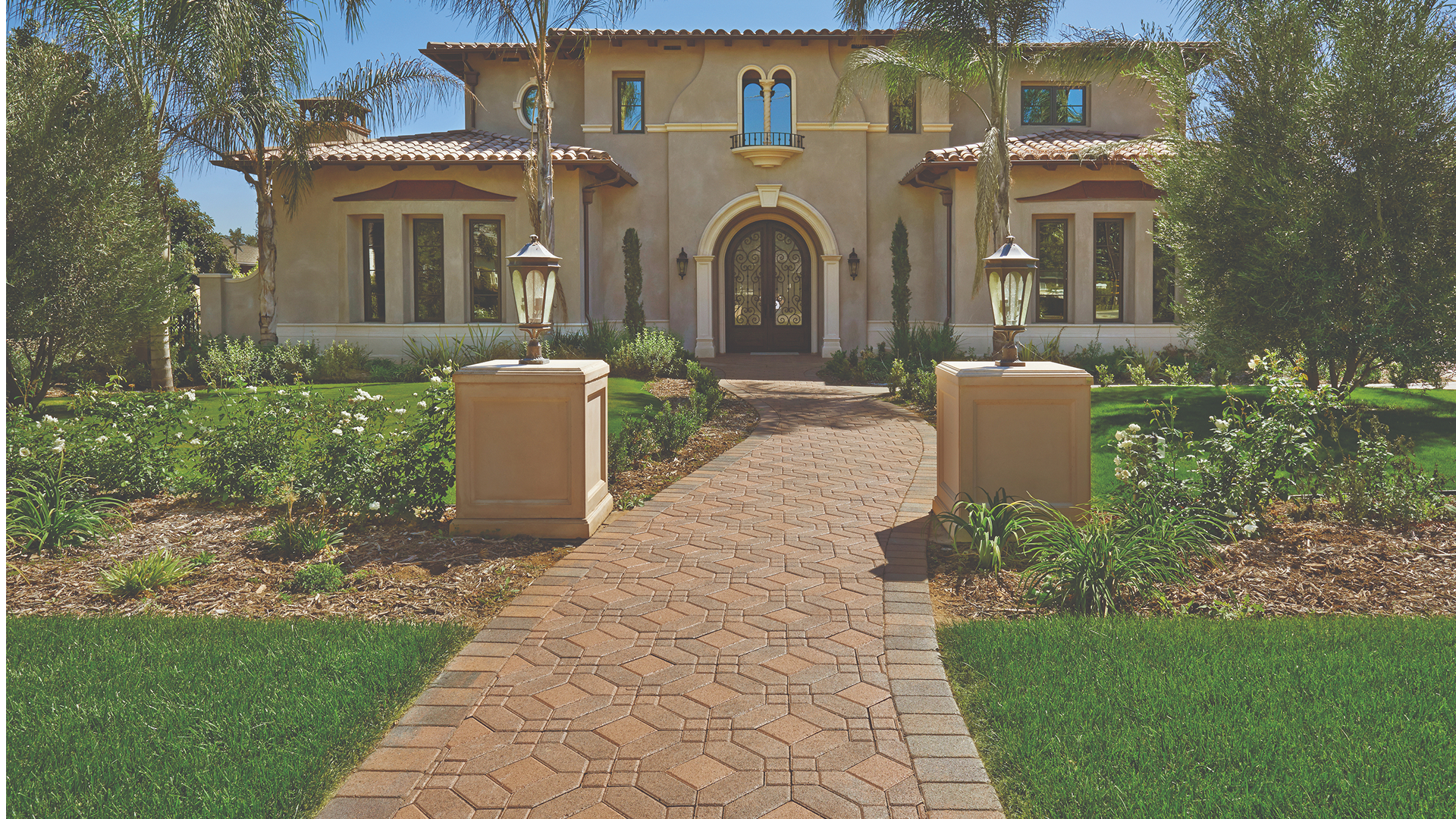 Cream-Brown Symetry® paving stones (special order color) combined with solid Cream 6x6 squares arranged in a Venetian parquet pattern. The elegant curved walkway is bordered with Tuscan Antique Cobble I and leads to a grand front entrance of a Mediterranean-style home framed by manicured landscaping and decorative lamp posts.